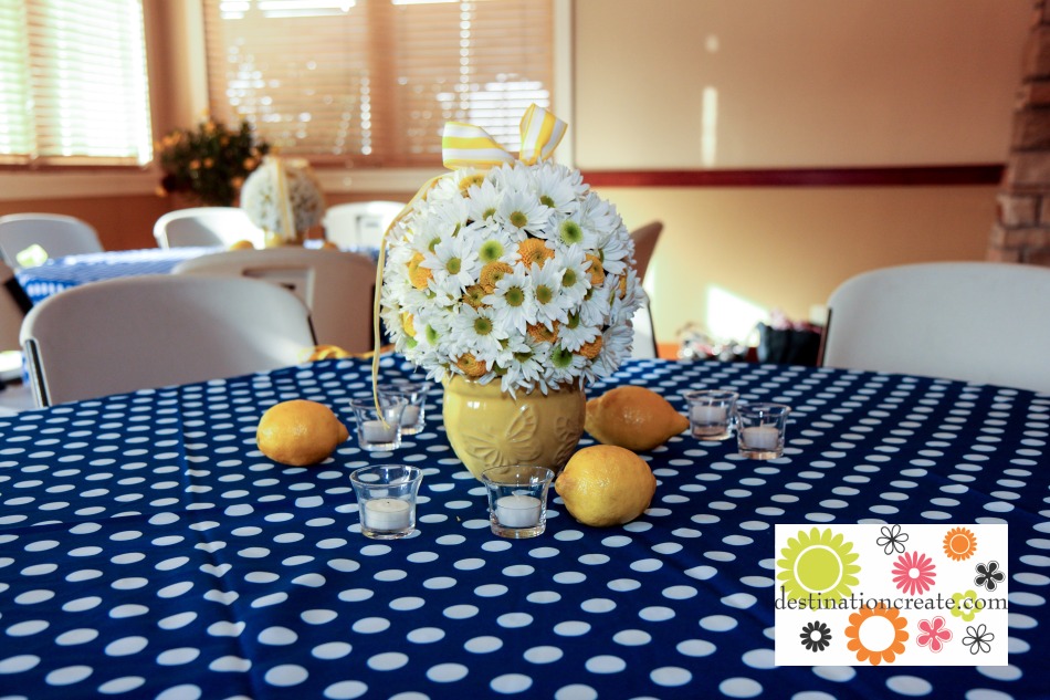 White daisy and yellow button mum flower ball centerpiece on yellow garden pot with lemons as table scatter.